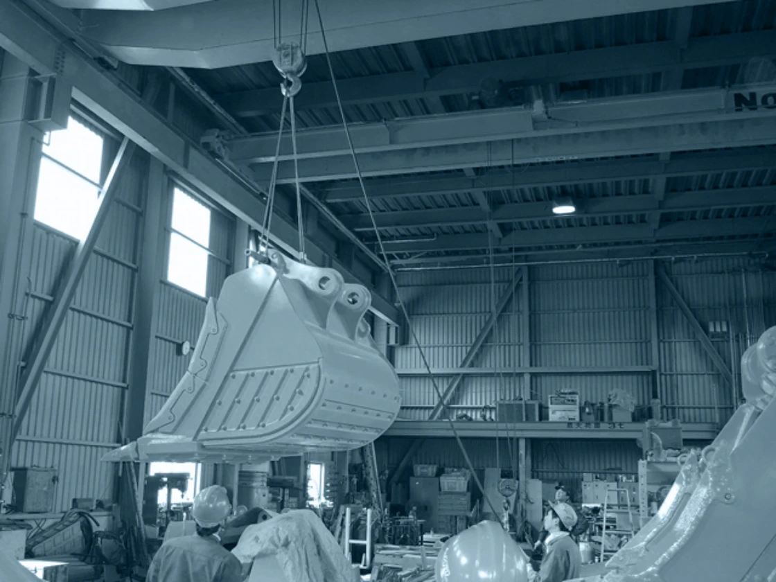 工場内でクレーンを操作する作業員と大型建設機械部品の吊り上げ作業。